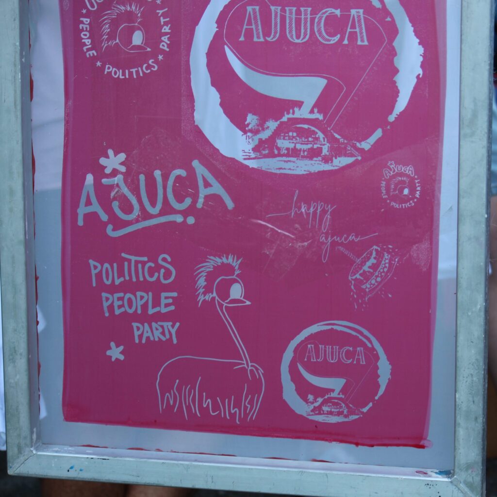 Ein grafisches Plakat mit bunten Schriftzügen zum "AJUCA" und die Worte ", "Politik", "People" und "Party" enthalten. Es zeigt auch eine einfache Zeichnung eines Vogels mit einem auffälligen Gefieder auf rosafarbenem Hintergrund.