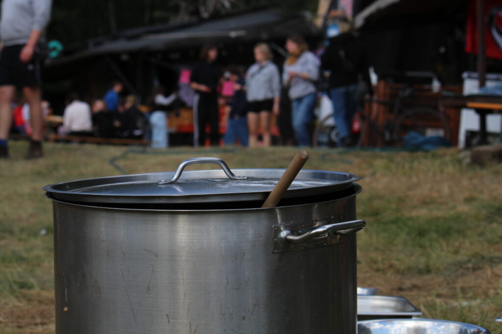 Ein großer Kochtopf mit einem Holzlöffel steht auf dem Boden, im Hintergrund sind Menschen und Bänke auf dem Alternative Jugendcamp sichtbar.